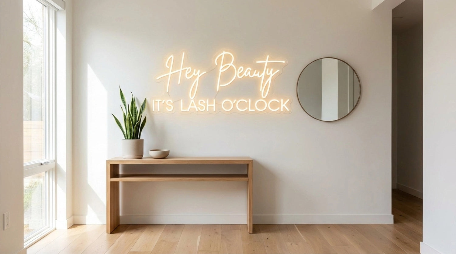 A modern and bright minimalist entryway featuring a custom MementoFun LED neon sign that reads "Hey Beauty IT'S LASH O'CLOCK" on a white wall. The scene includes a light-wood console table with a potted snake plant, a round wall mirror, and natural sunlight, showcasing a perfect personalized housewarming gift idea.